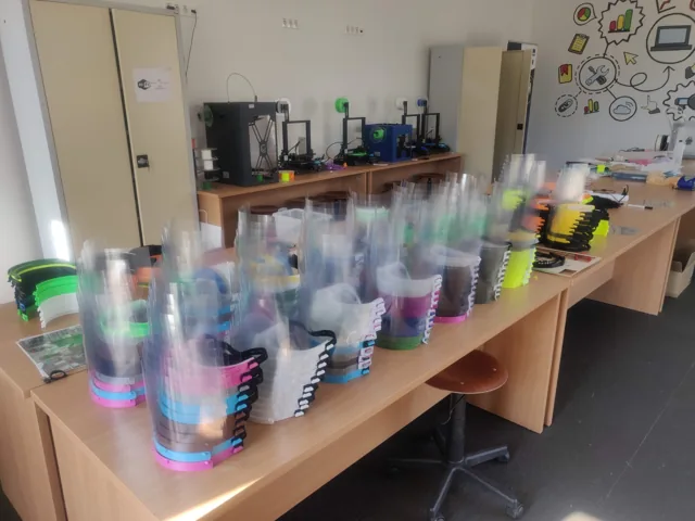 Stacks of finished face shields in every filament color on a long table, the printers that made them along the back wall